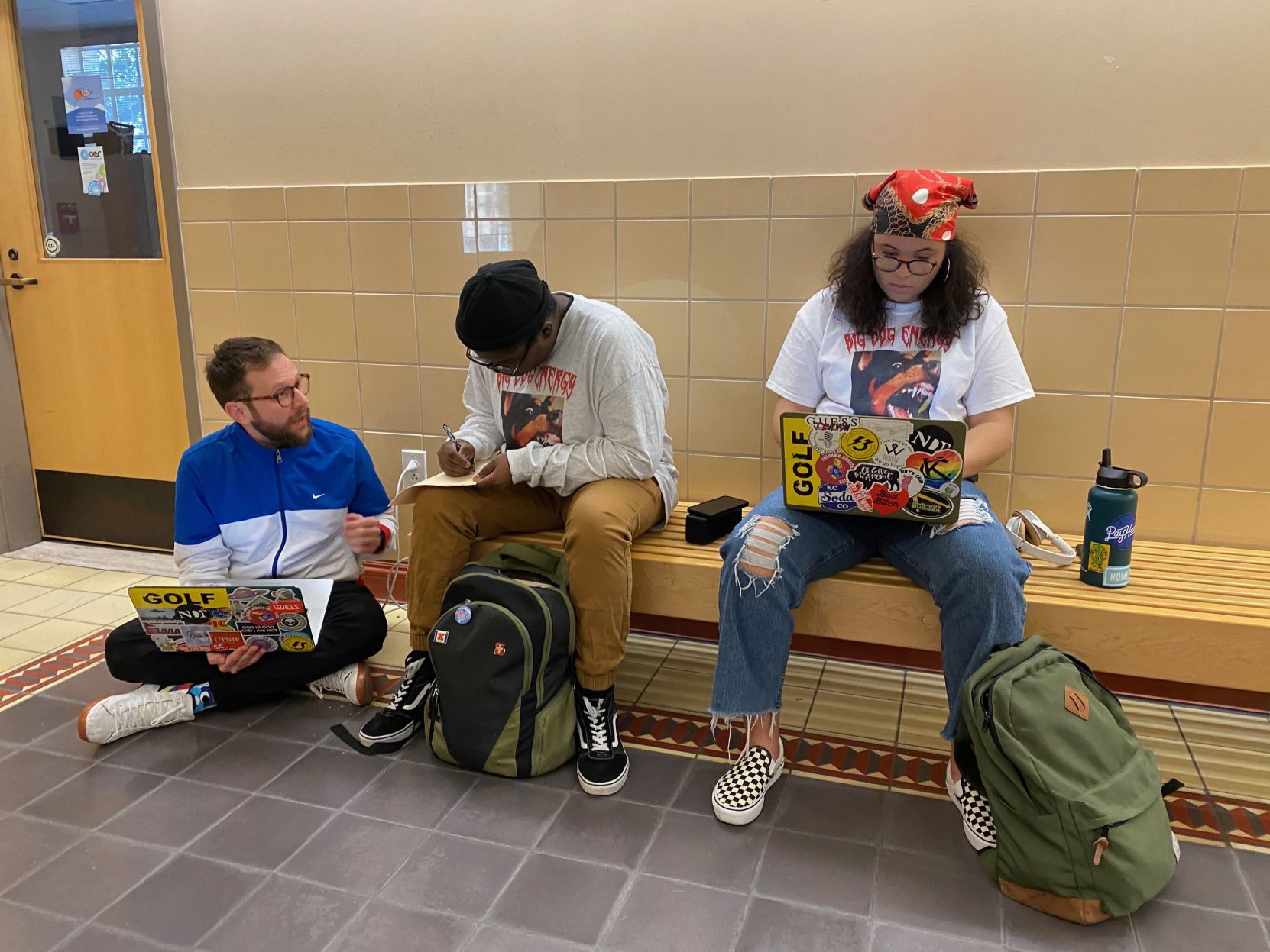 Debate coach Sean Kennedy sits on the ground next to two debaters on a bench working on a bench.
